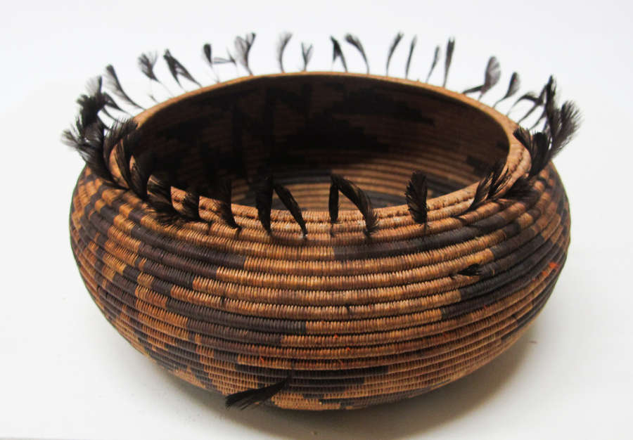 Woven tan and brown onion shaped bowl with geometric dark brown patterning and feather-like tassels emerging from the top edge of the bowl.