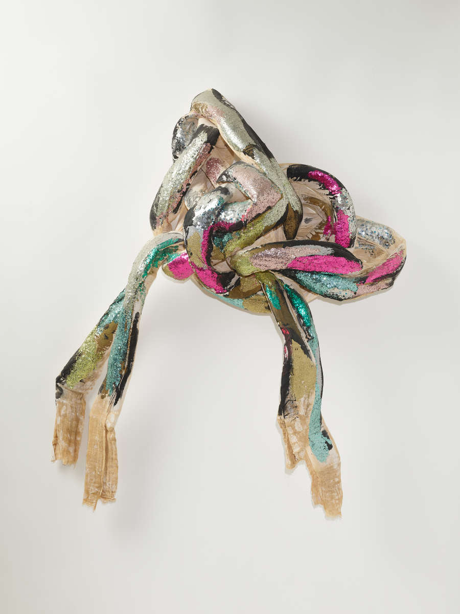A knotted bundle of textile strips with metallic silver, pink, green, and black cloth, with long frayed ends hanging down loosely. 