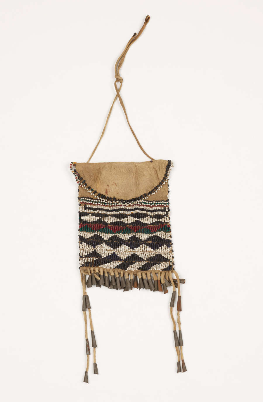 A leather beaded pouch with white, black, red, and green beaded designs. There is a handle as well as fringes with metal and leather decorations.
