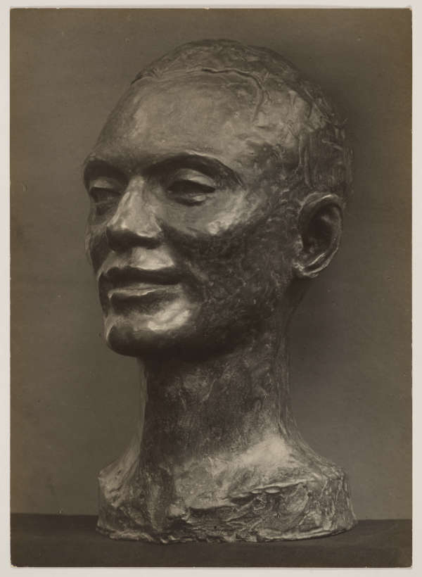 Photograph depicting a textured, bronze sculpture of the head of a male presenting figure; presented in a three quarter view against a dark backdrop. The face features a slight smile.