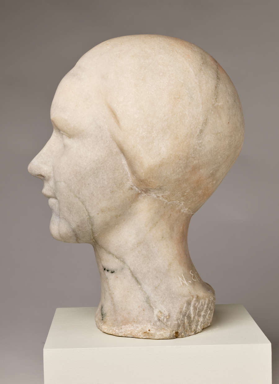 Profile view of a blemished marble sculpture depicting the head of a female figure. The face features a subtle expression and delicate details, framed by a tight hairstyle.