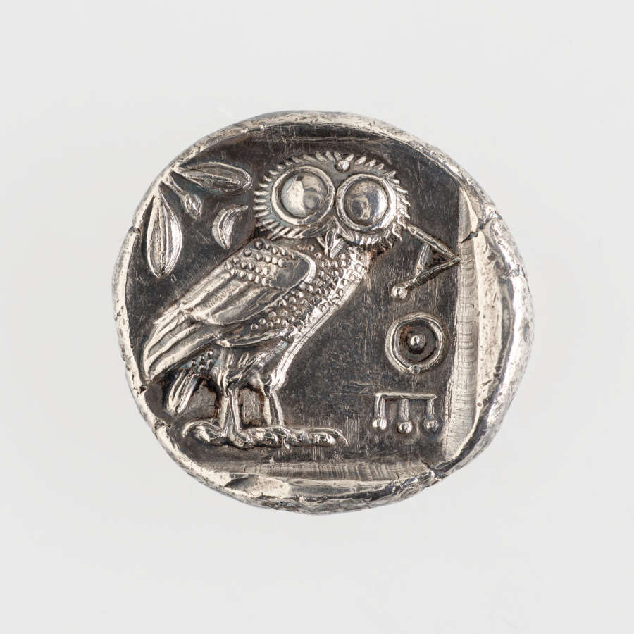 Back of an irregularly round silver coin, embossed with an uncentered image of foliage, the Athenian owl, and the Greek letters “AOE” beside it.