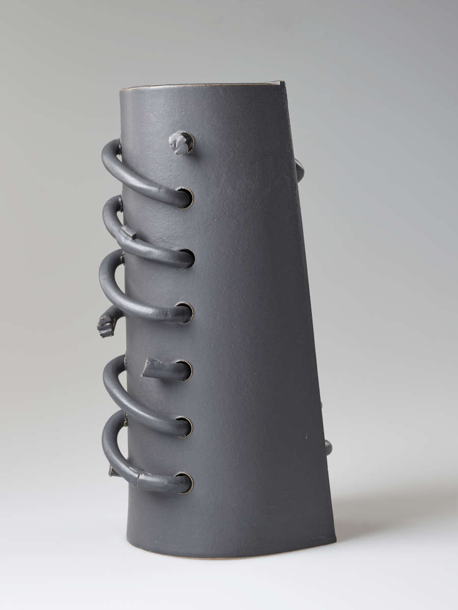 Diagonal back view of a cylindrical gray sculpture featuring a row of helical protruding loops threaded through the ceramic surface. Some of the loops are broken.