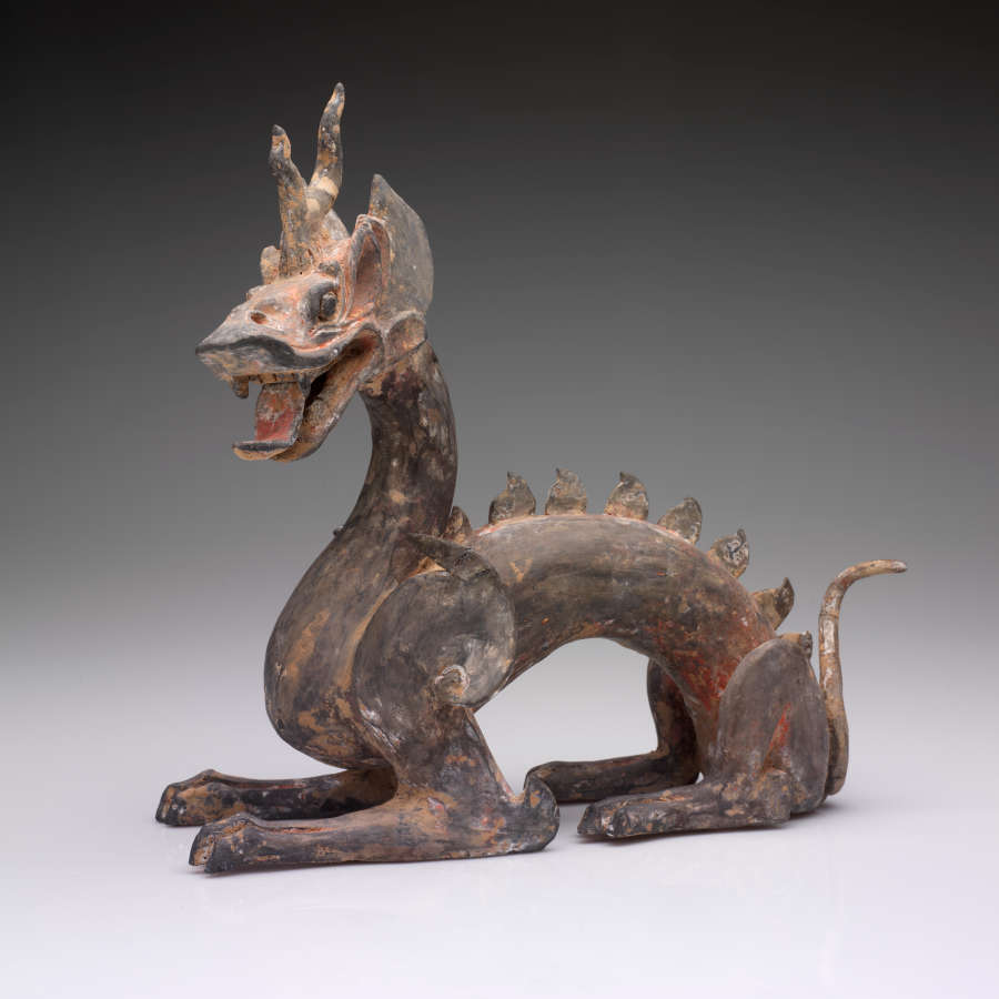 Side-view of an arched four-legged dragon with a lean body, open mouth, horned head, scaly back and a thin tail. The paint has worn into patches of browns and reds.