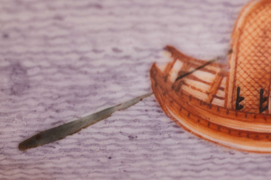 A close-up of a painted scene on a ceramic bowl depicting a small wooden boat with a woven canopy, a single oar extending off the side into purple-hued water.