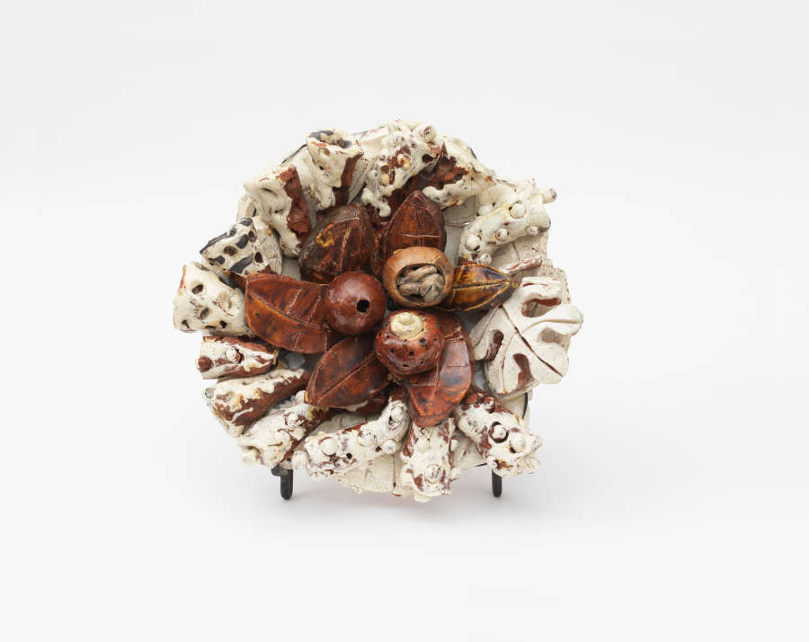 Circular ceramic sculpture featuring a cluster of glossy brown, seed-like pods at the center, surrounded by textured cream leaves speckled with brown radiating outwards from the cluster of seeds.