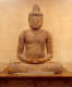 A weathered wooden Buddha statue sitting cross-legged on a wooden platform, with hands resting in the lap, with a serene expression and closed eyes. 