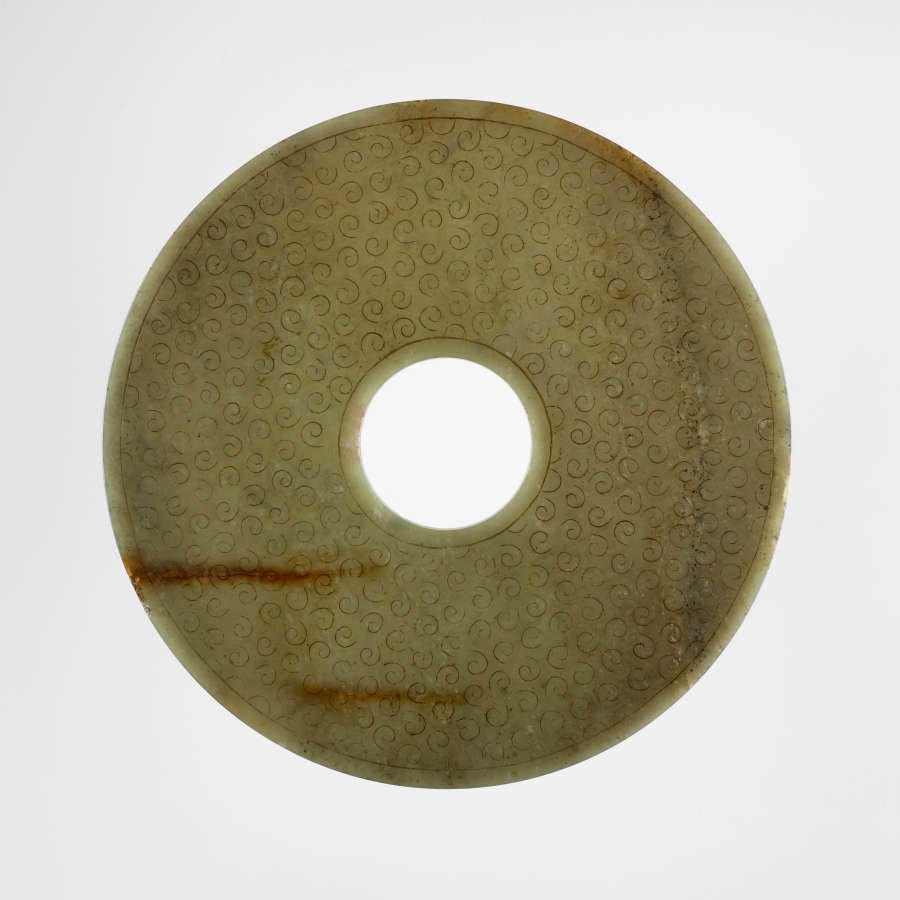 The other side of the circular object, which features small swirling patterns engraved onto a muted-green surface with two blurry brown streaks. 