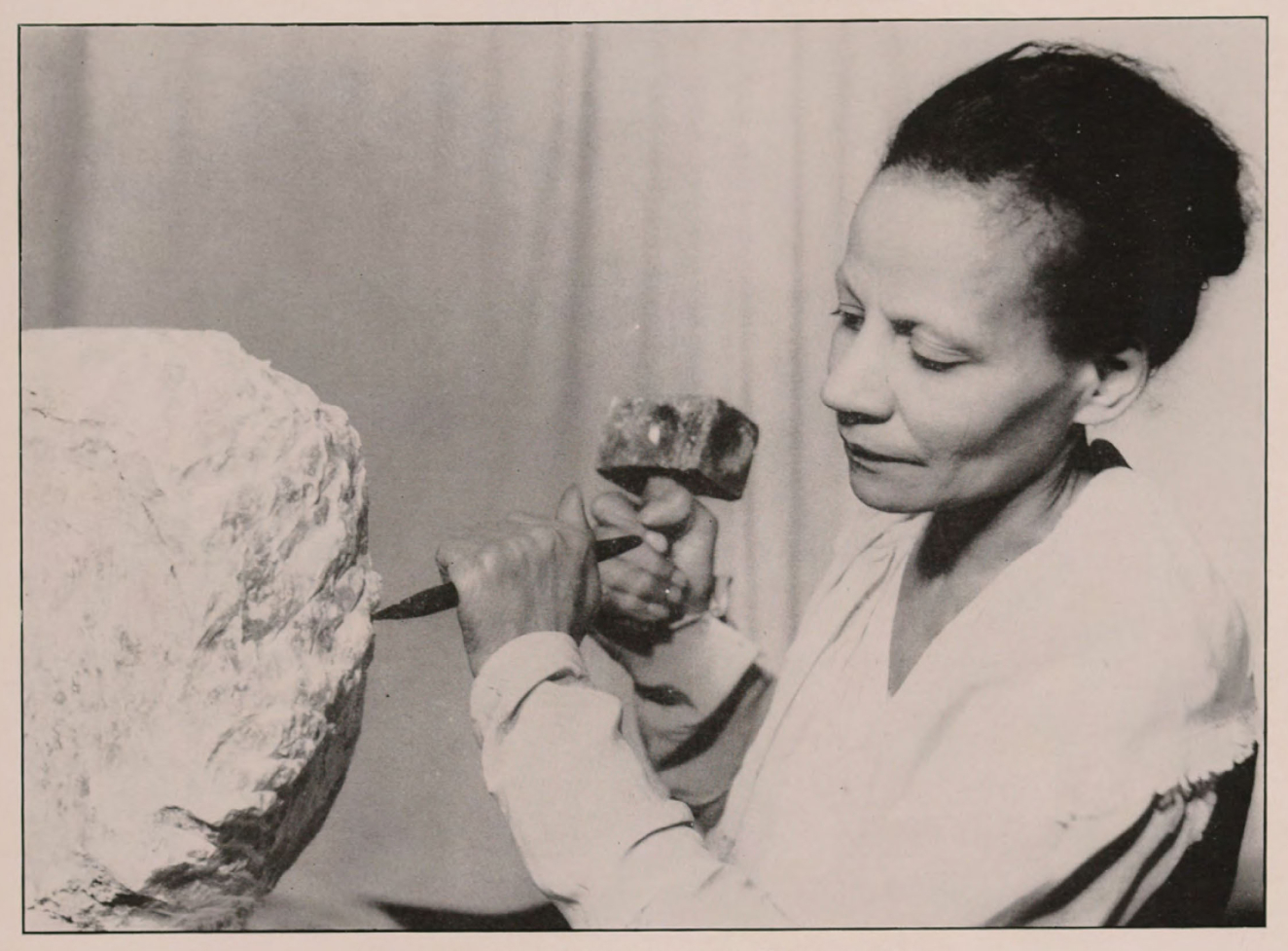 Black and white photograph of a woman of color sculpting a large stone block with hammer and chisel in hand. She is intent on her task. 