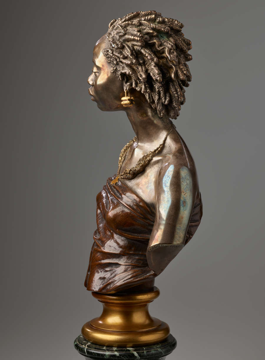 Profile of a bronze bust of a woman, hair in twists swept back, gold earrings, beaded necklaces, and shoulders exposed in a dark brown wrapped top mounted on a gold base.
