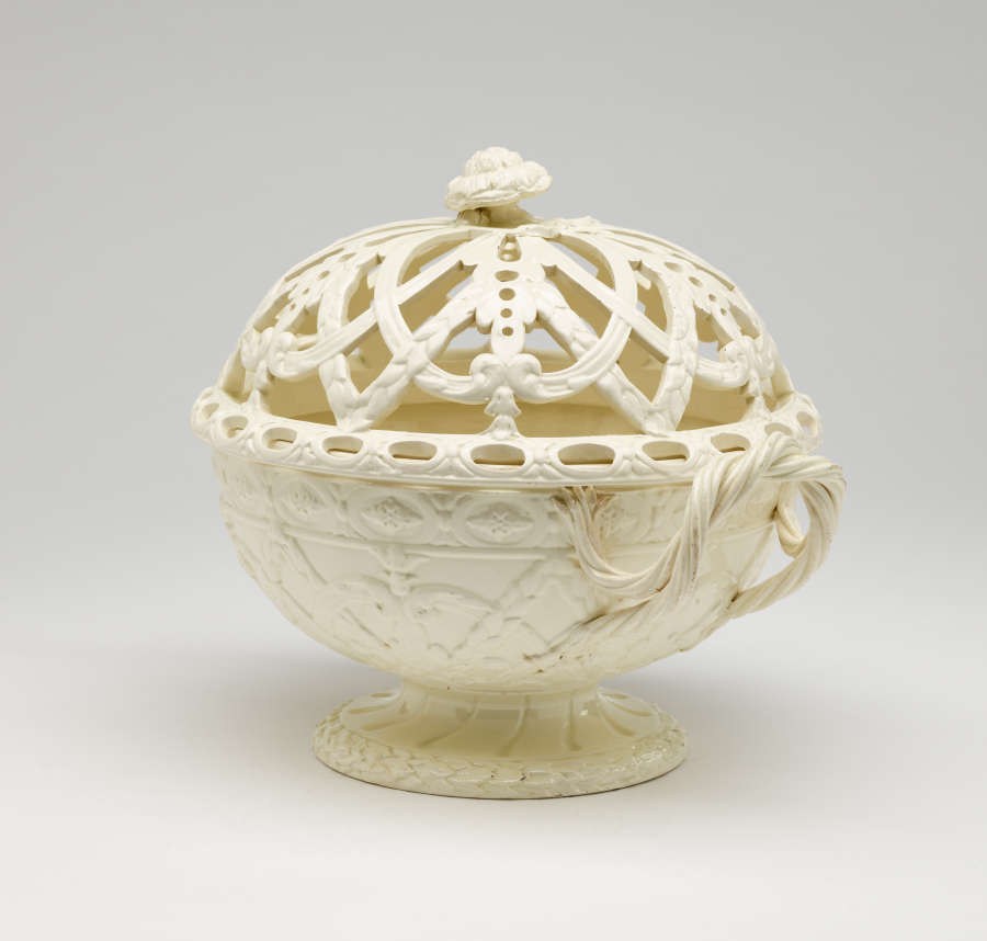 A cream-colored basket with sculptural woven handles, sculpted foot, and a lid with cutout designs.