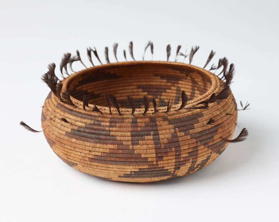 Woven tan and brown onion shaped bowl with geometric dark brown patterning and feather-like tassels emerging from the top edge of the bowl.