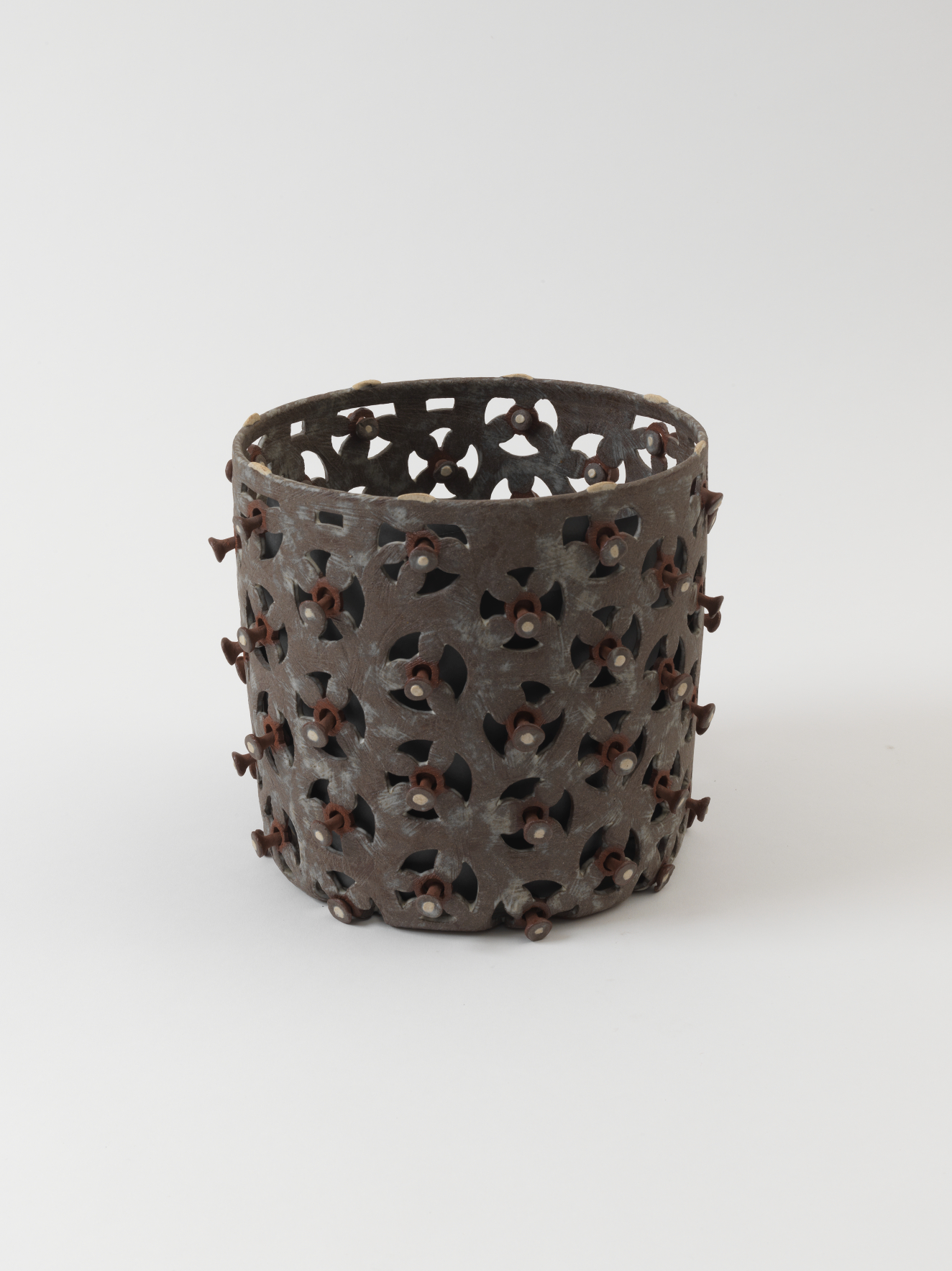 Dark brown cylindrical vessel with evenly spaced repeating triangular cutouts that form irregular circles. The center of each circle is punctuated by a raised round stud.