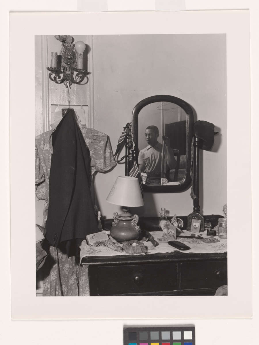 A grayscale photograph of a vanity covered in memorabilia. The mirror on the vanity reveals a dark-skinned figure in the room, holding a crutch in one hand.
