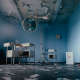 Photograph of an abandoned-looking operating room with chipping ceilings, blue walls, and discarded paper and debris scattering the gray floor. Aged medical equipment and white metal furniture splay the room.
