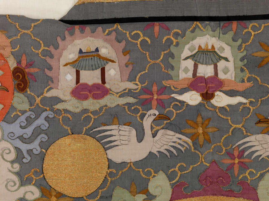 Robe’s back detail. Two flaming and haloed pagodas on clouds are above a golden circle and white bird, amongst earthy pastel clouds and a dark background with a diagonal grid.