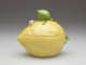 Lidded yellow box shaped like a lemon. Molded green leaves with a large bud and small pink flowers are applied to the top and wrap around the side.
