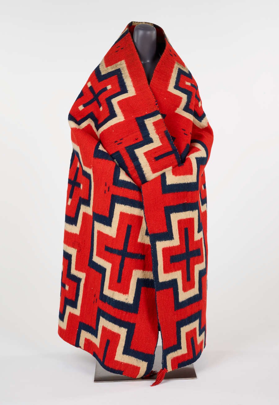 Photo of a large, red woven blanket, with geometric cross-shaped patterns, wrapped around a headless mannequin. The blanket’s red tassels hang when the blanket is worn as a garment.