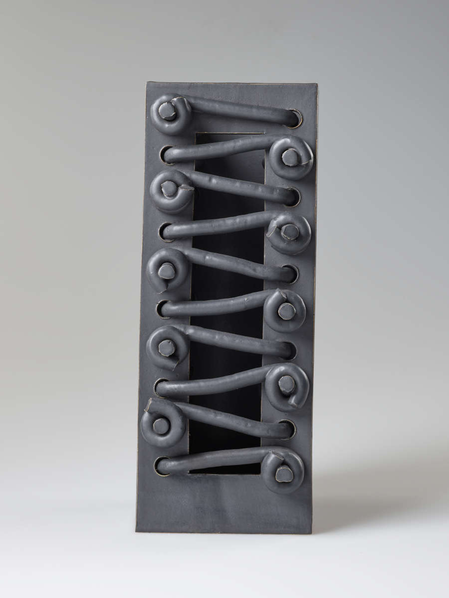  Front view of an abstract, gray, industrial-like, rectangular ceramic form. A row of knots are laced across sides of the front flat periphery with a centered rectangular cavity in between.