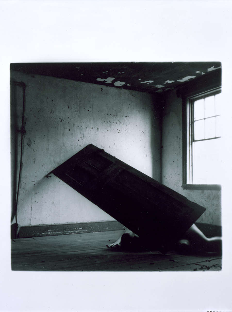 Dreamy black and white photo of a door propped precariously against a wall. A figure crouches beneath, a pale arm and leg visible. Light streams in a window at left.