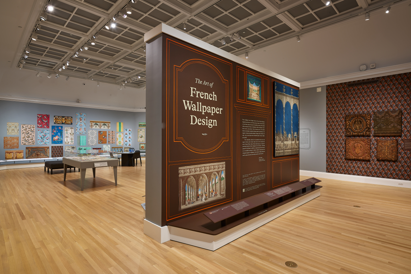 A large brown wall titled “The Art of French Wallpaper Design” introduces an exhibition of multi-colored, decorative wallpaper samples arranged within the open gallery space behind it. 