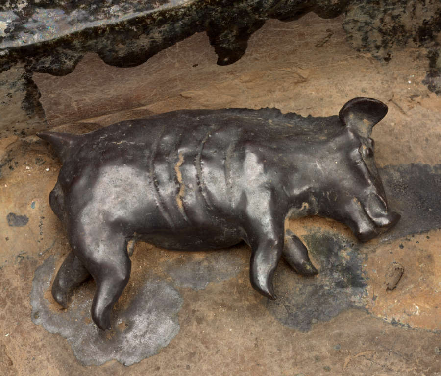 Detail of the ceramic sculpture, showing a small pic laying on its side in the enclosure. The pig’s surface is dark and glossy, its body bearing ridges resembling large scars.