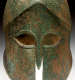 Bronze helmet with a corroded surface and green discoloration. Features large sharp eye holes with carved insets connected to a narrow nose guard in the helmet's center.

