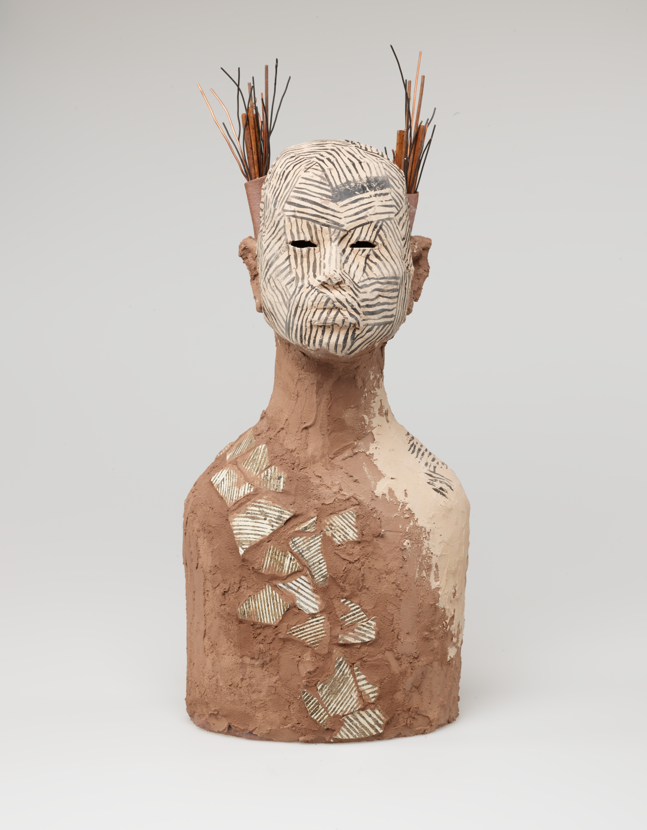 Brown clay bust of a figure with a grass and stick headdress. Diagonal lines of patterned patches of offwhite run across their chest and cover their face.