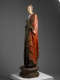 A tall, weathered wooden statue of a robed figure with a serene expression, draped in green and red garments, standing upon an octagonal base.