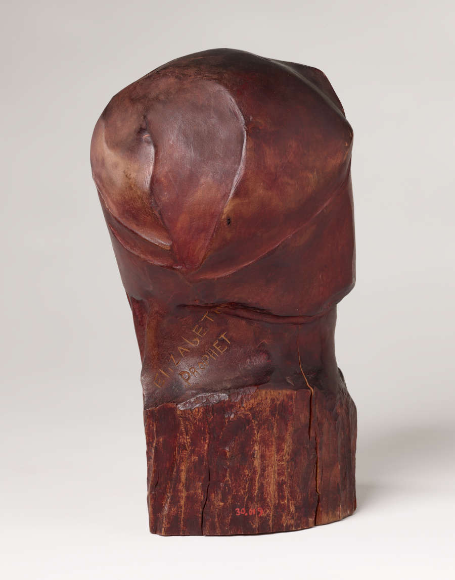 Back view of a carved wood sculpture depicting a head and neck. It is smooth, besides the bottom, where the wood is left uncarved and features cracks. The signature “Elizabeth Prophet” is thinly carved into the neck.