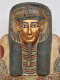 A gilded face with blue-outlined eyes on a coffin lid, framed by a blue black headdress with golden details and a red, green, and blue painted collar.