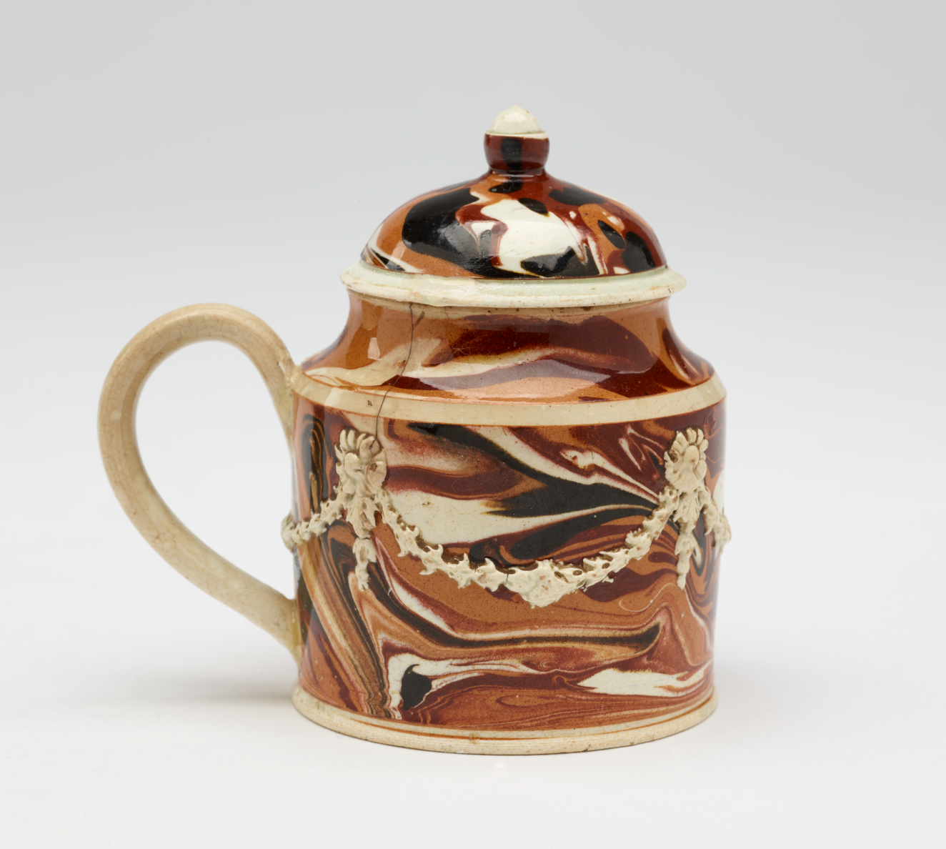 A small mustard pot that is white, light brown and dark brown. The lid and handle are rounded and there are swag floral decorations on the body.