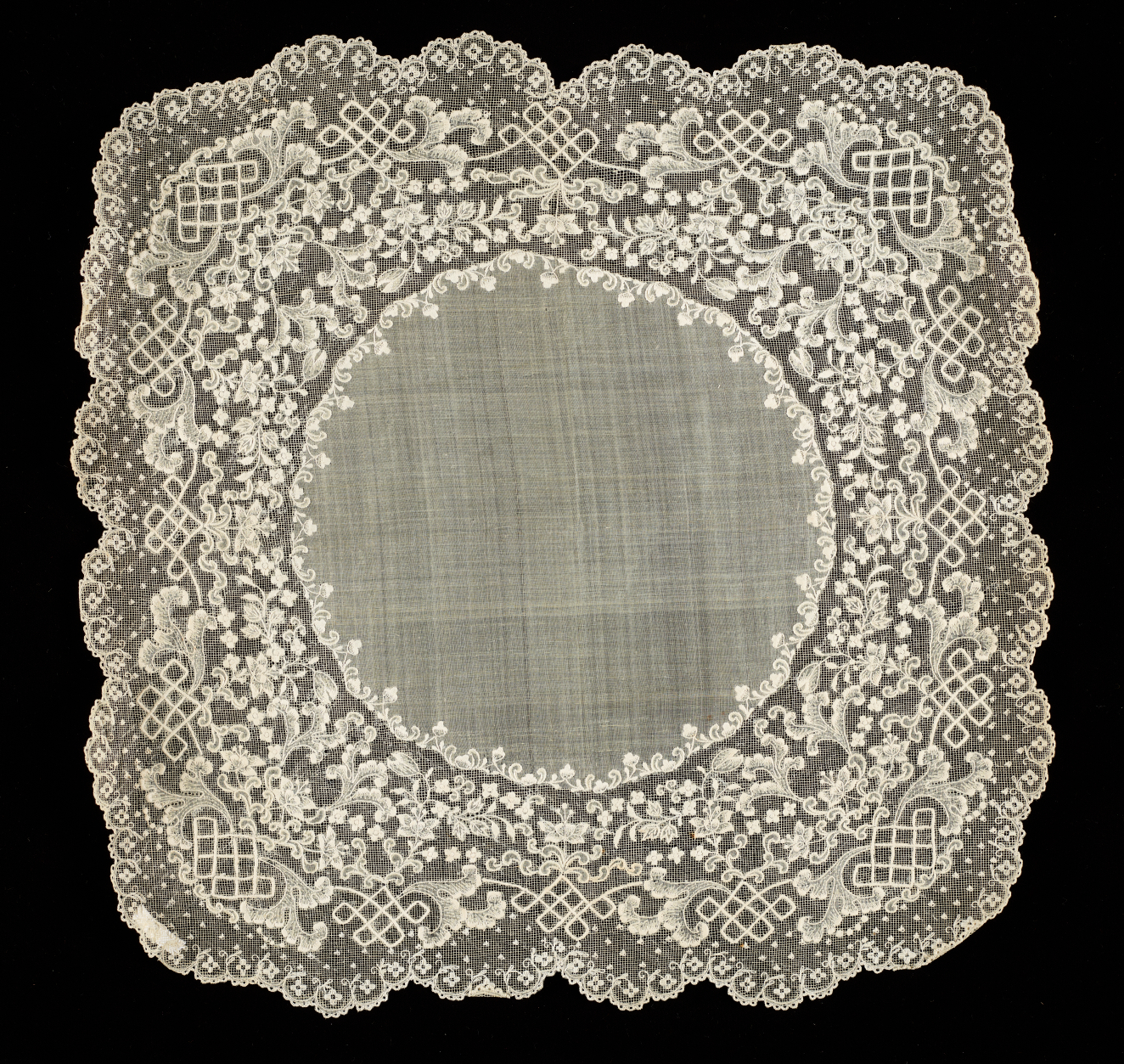 Square of fabric with rounded edges and a lacy border. The surface features intricate embroidery depicting intertwined curving lattices around delicate flowers surrounding a circle of plain fabric.