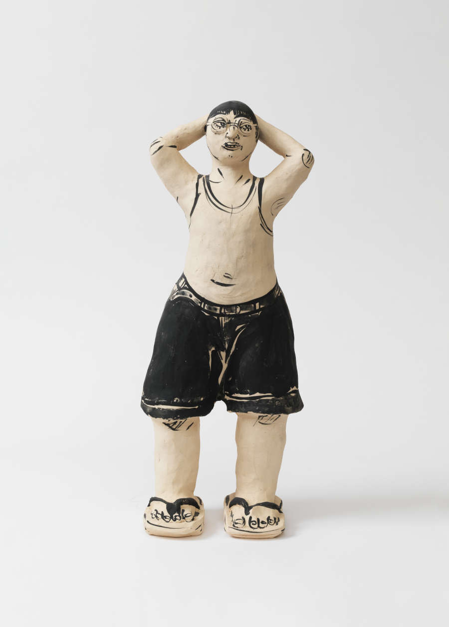 Ceramic sculpture of a boy standing upright with hands clasped behind his head, wearing black shorts, tank top, and sandals, his body rendered in solid black-and-white and stylized outlines.