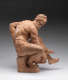 Realistically carved terracotta sculpture depicting a muscular bearded man sitting facing right,  one arm bent behind him in support as he leans over to hold the vessel between his legs.