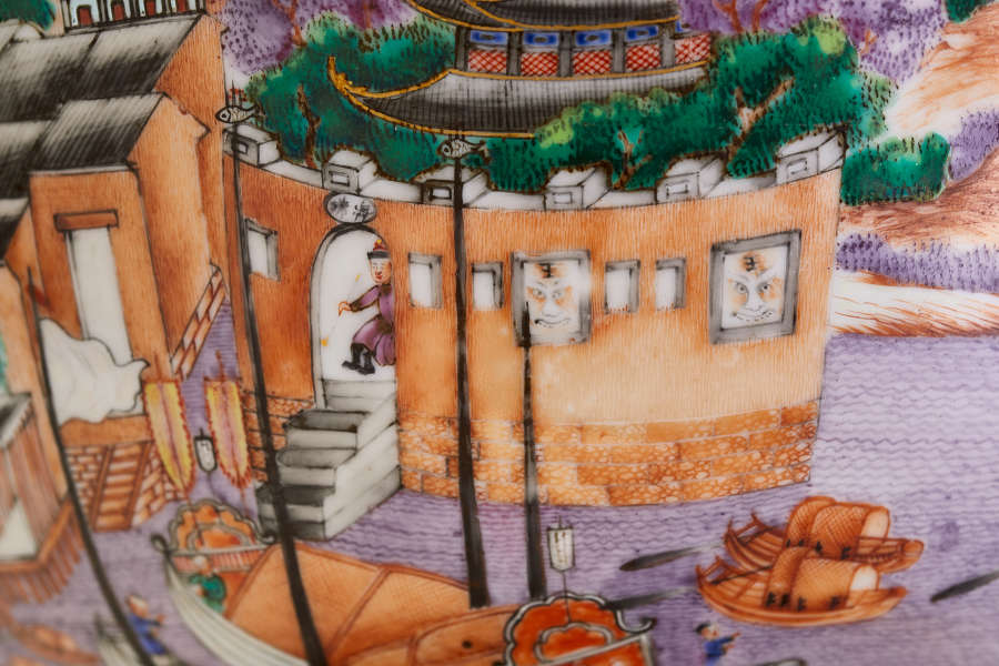 A close up of a painted ceramic featuring an orange walled structure with rectangular windows, a figure in purples robes exiting through an arched doorway, surrounded by boats and greenery.