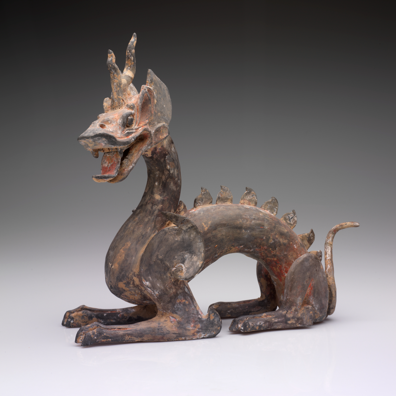 Side-view of an arched four-legged dragon with a lean body, open mouth, horned head, scaly back and a thin tail. The paint has worn into patches of browns and reds.