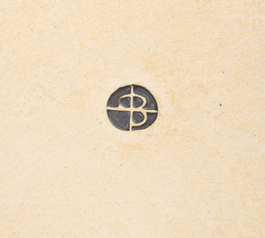 A small imprint on a beige ceramic surface. It features a decal that resembles an overlaid and reflected “P” and “B” enclosed in a circle and filled with black glaze.
