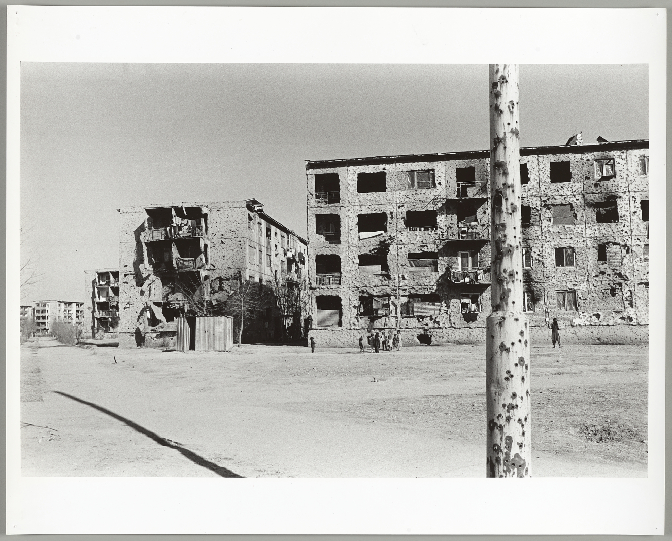 A black-and-white scene of a bullet-riddled pole in the foreground and heavily damaged buildings with crumbling walls and blown out windows in the background. 
