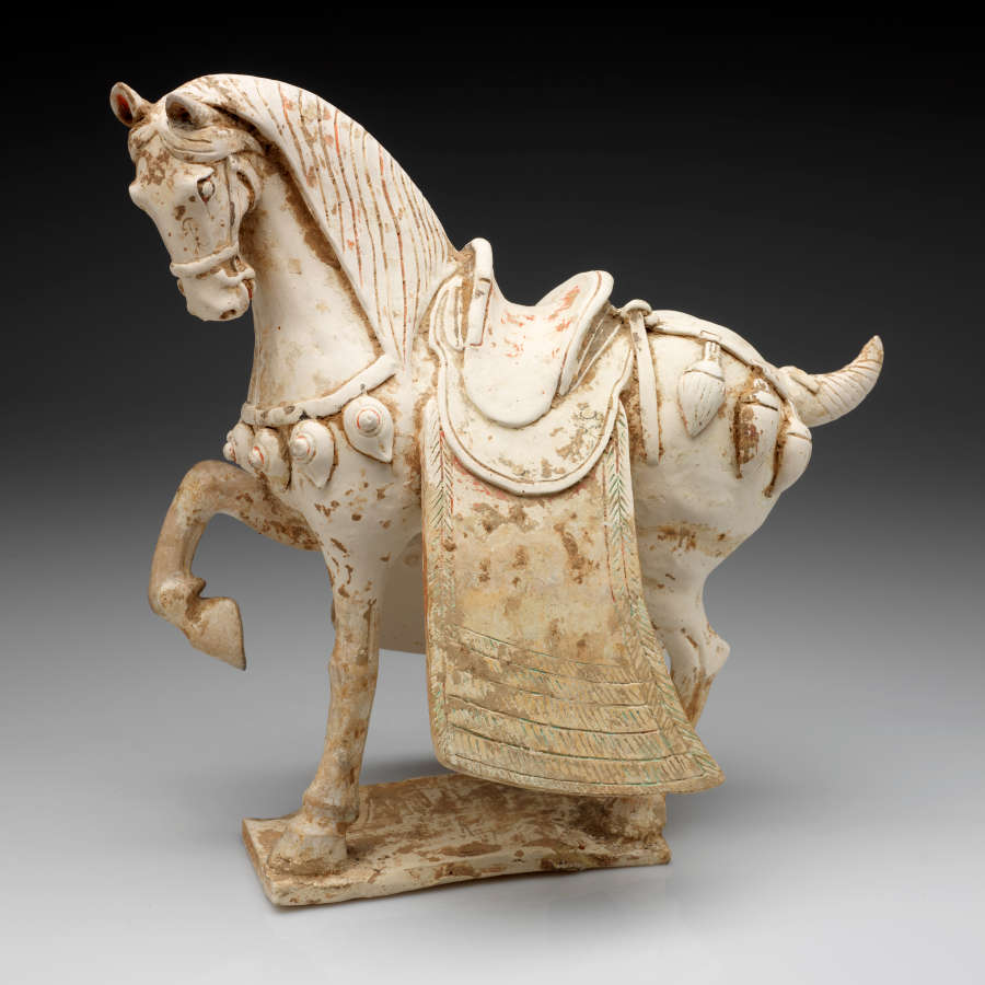 Side-view of a white ceramic sculpture of a saddled ornamented horse with one foot raised, a short tail, a heavy mane, and facing leftwards. It stands on a thin base.