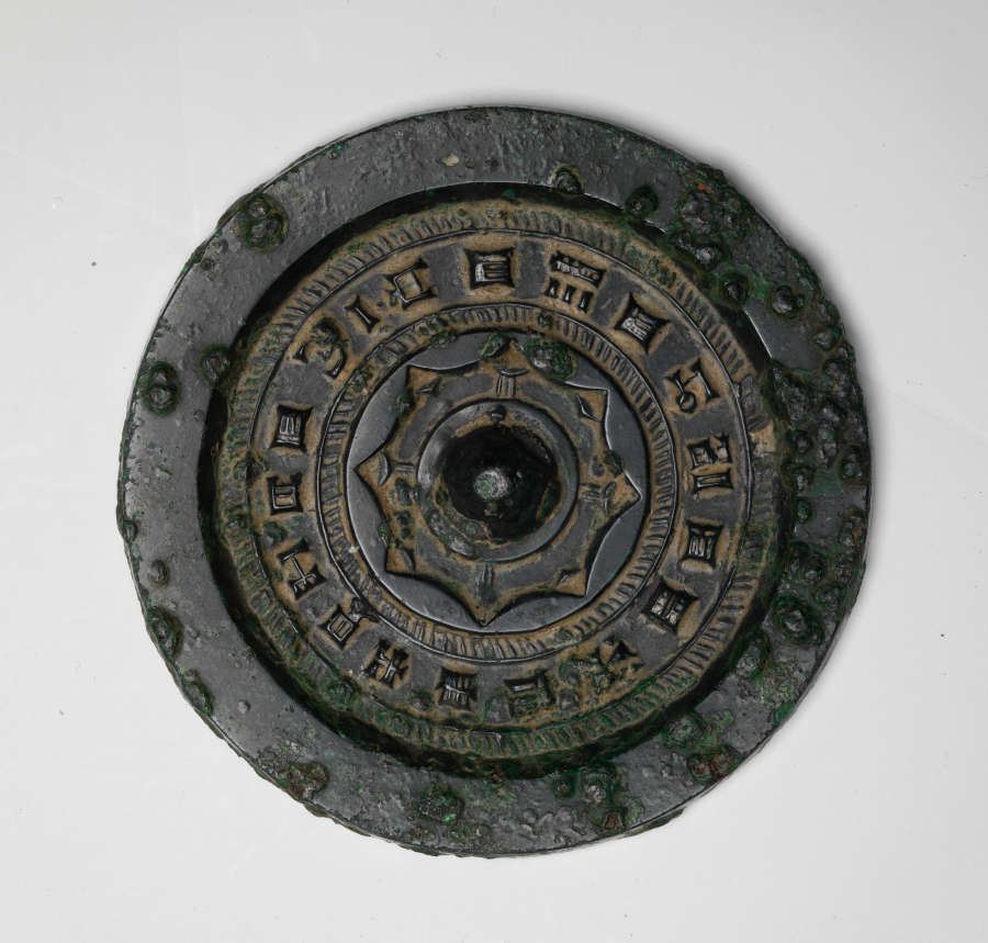 Back of a circular corroded bronze mirror, embellished with a thick raised border, concentric engraved patterns, writing and a domed center. Surrounding the raised writing are yellow stains.