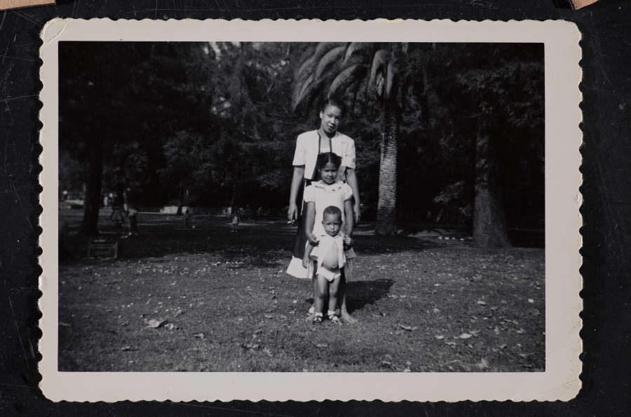  Snapshot of a woman under a palm tree, a girl and toddler standing directly in front of her. All have medium-dark skin. The toddler’s open shirt reveals his round tummy.