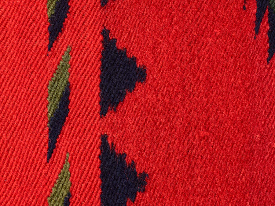 Closeup of a red tapestry with alternating woven textures. The tapestry has a blue, jagged trim and geometric patterns. Next to the triangles are blue-black and green vertical rhombuses. 