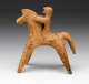 Side view of a terracotta figurine of the simplified forms of a horse with rider, decorated with faded red painted lines. The figure sits upright, clinging to the horse’s neck.