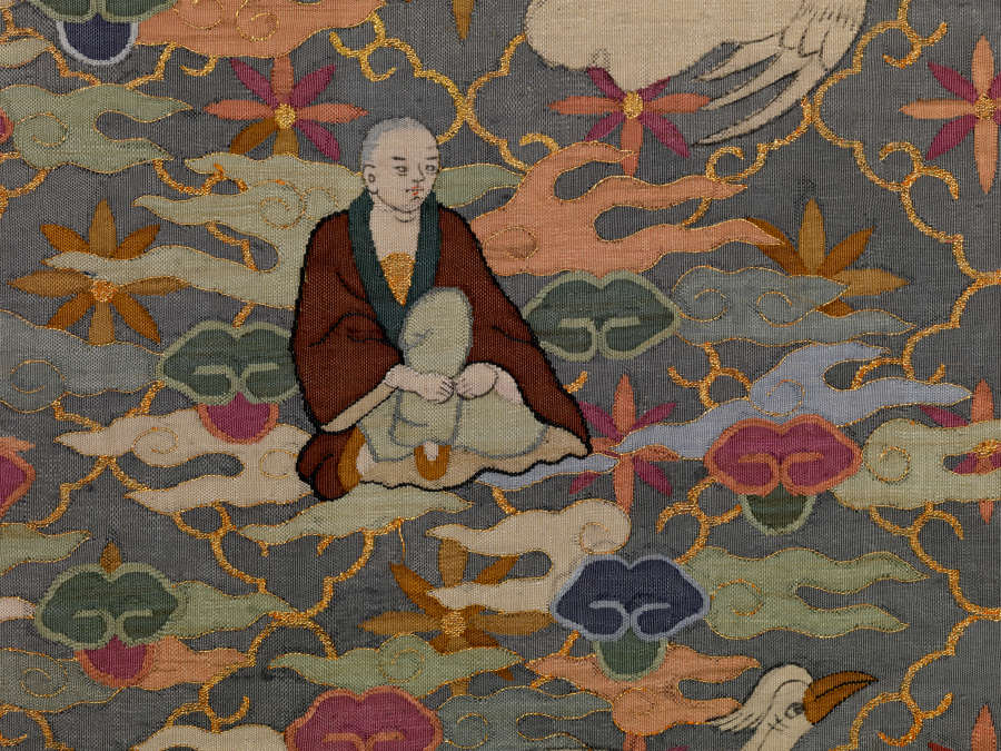 Section of illustrations on the blue robes back, featuring an intricate floral pattern overlaid with densely-packed pastel wispy clouds, a flying white bird and a seated monk in brown robes.