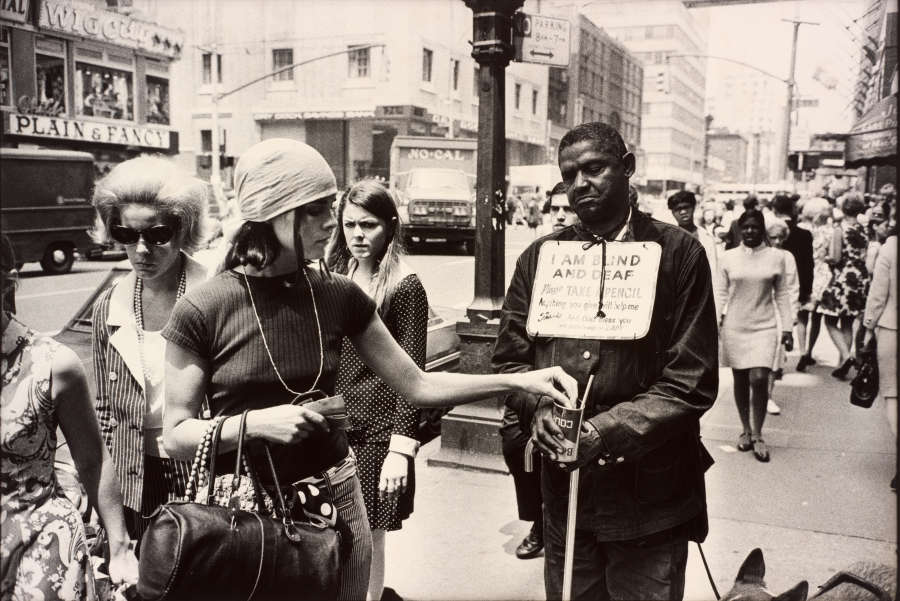 Black-and-white crowded street scene showing a dark-skinned person wearing a sign reading “I am blind and deaf” and a fair-skinned young person dropping a donation into his cup while another fair-skinned onlooker stares in disgust. 