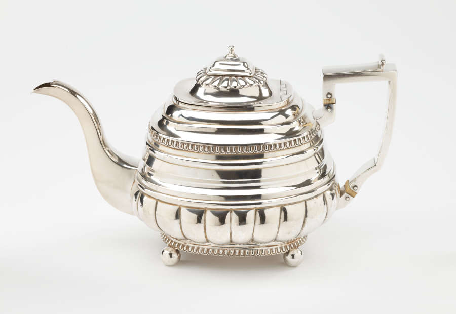 A silver teapot with a decorative angular handle, a rounded square body and spout with sculptural decorations.