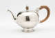A bulbous silver teapot with a wooden finial and handle connected with decorative silver pieces. There are delicate engraved marks on the body and lid.