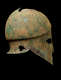 Bronze helmet with a narrow nose guard and sharp eye holes in profile facing right. The corroded surface is brown with significant green discoloration. The top has jagged sections missing along the crown.