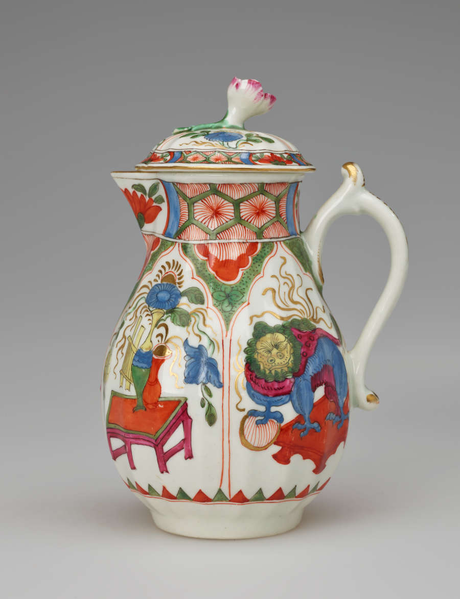 A white milk jug with a handle, lid, and spout with green, red, blue, pink, yellow, gilded decorations,and two vignettes; an animal and a table, vessels, and floral elements.